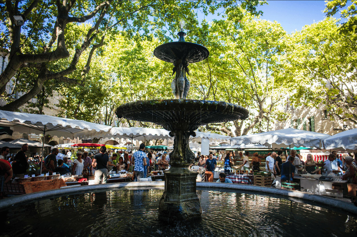 Scène du marché hebdomadaire d’Uzès sur la Place aux Herbes, vue sur la fontaine et des gens qui se baladent entre les étals