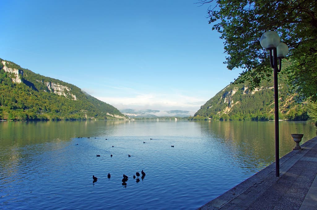Lac de Nantua sous un ciel bleu entouré de montagnes verdoyantes de chaque côté.