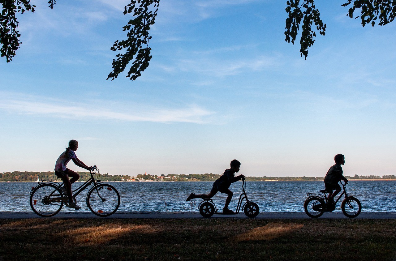 Silhouettes d'enfants et d'un adulte à vélo et trottinette, longeant un lac au coucher du soleil.