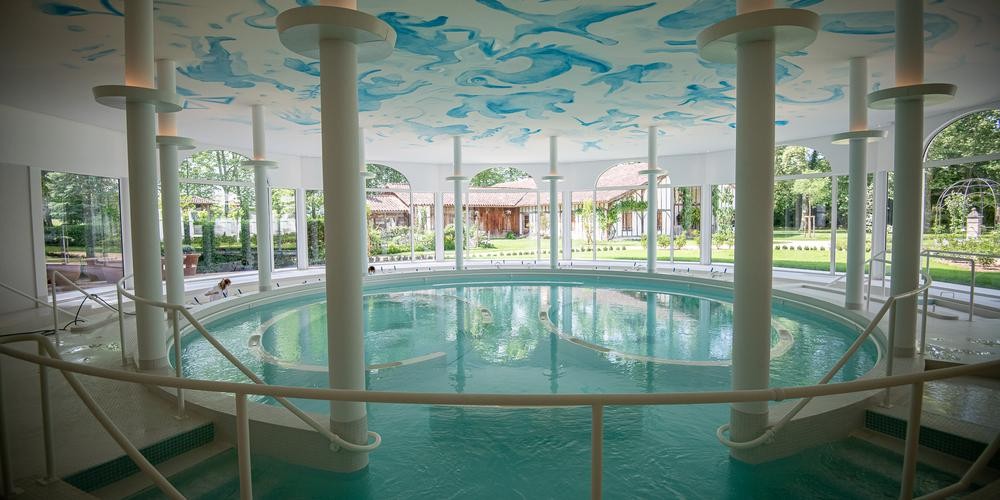 Piscine intérieure circulaire avec colonnes blanches, entourée de baies vitrées offrant une vue sur un jardin verdoyant.