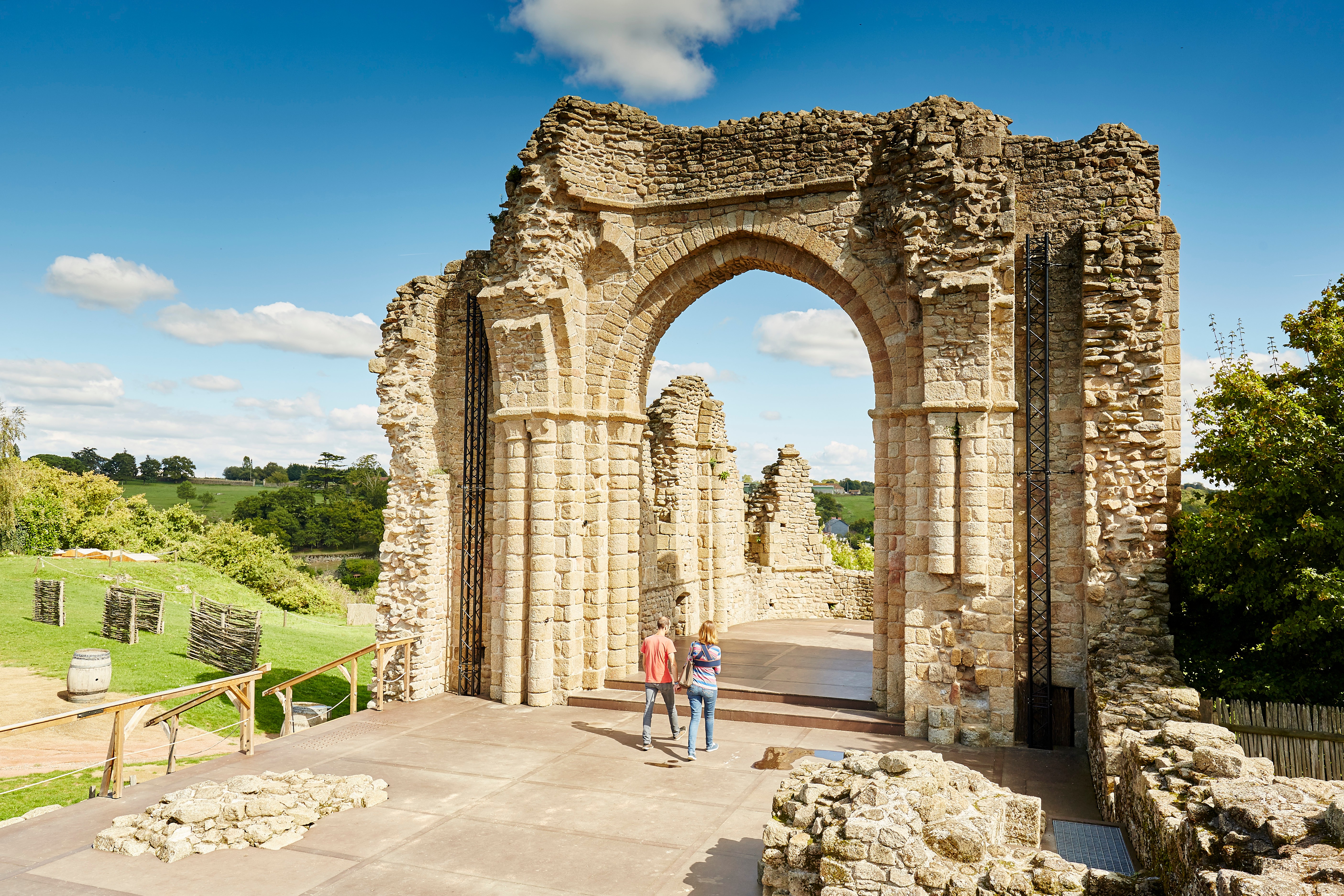 Vue sur des vestiges en pierres sur un sol carrelé entourés de verdures et 2 personnes entrant par une arche.