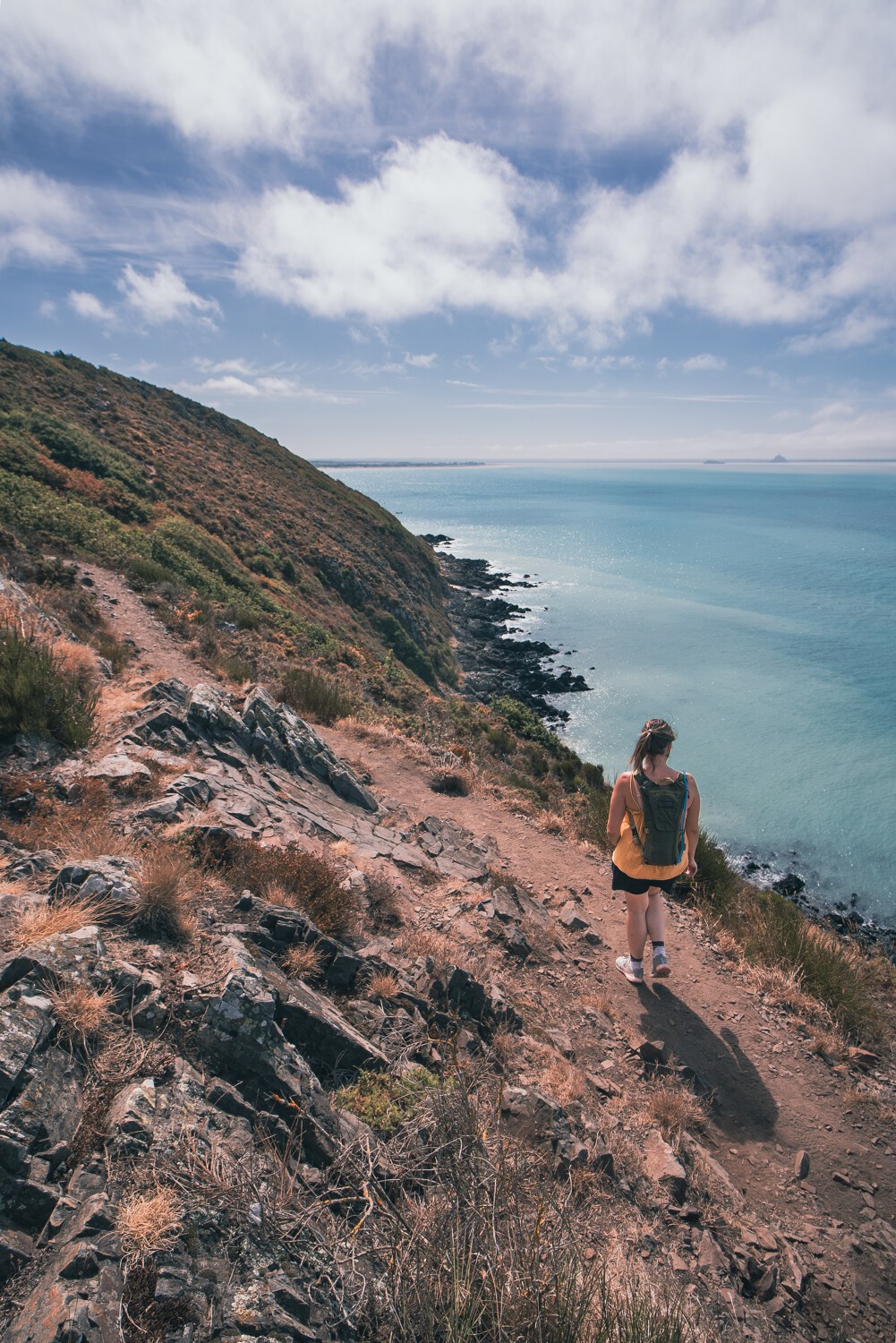 Randonneuse, GR223, pierres, chemin, falaise au-dessus de la mer bleue, ciel bleu nuageux 