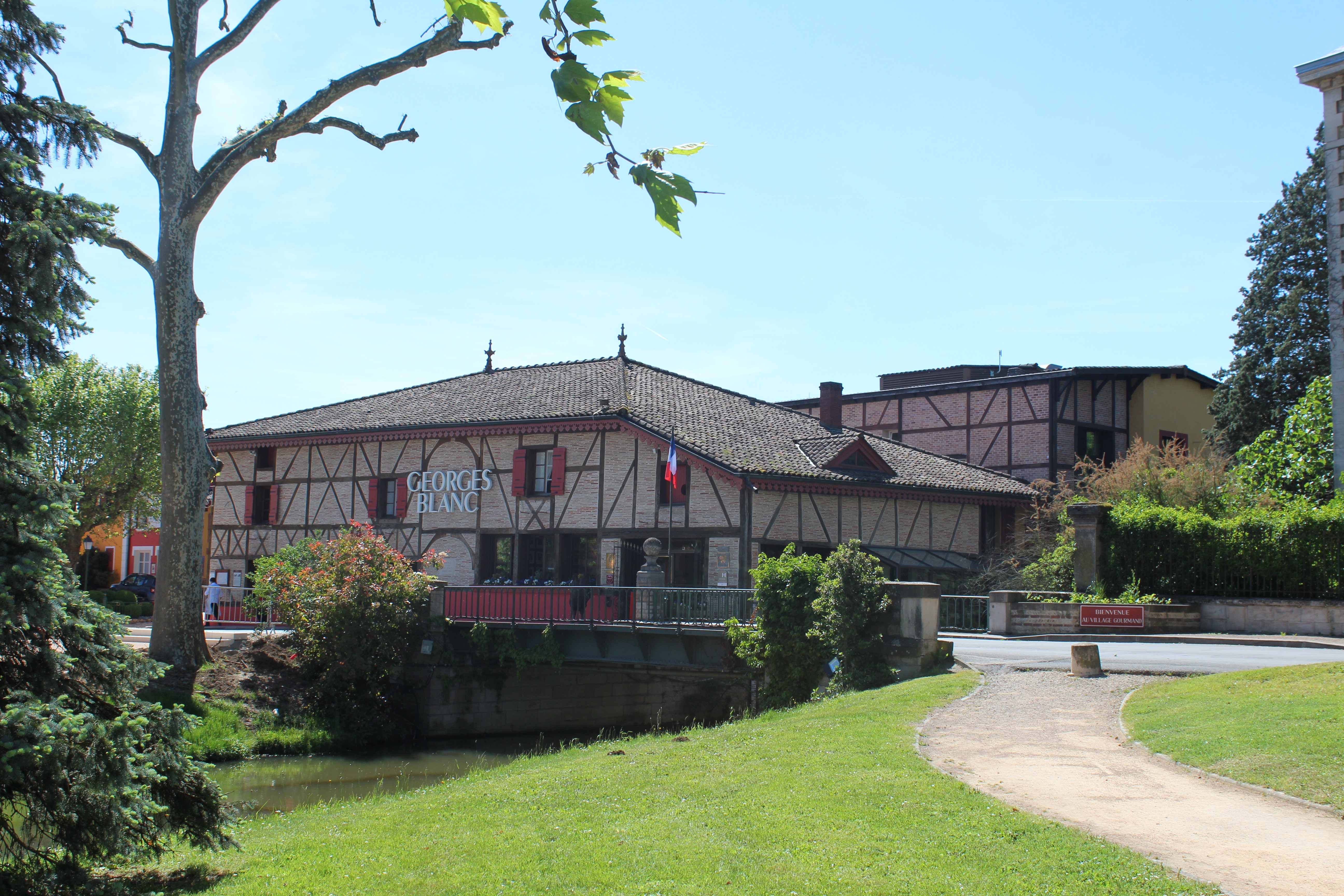 Devanture du restaurant “George Blanc”, dans son architecture bressanes avec des petites briques, poutres apparentes. En premier plan un sentier de terre au milieu de verdure.