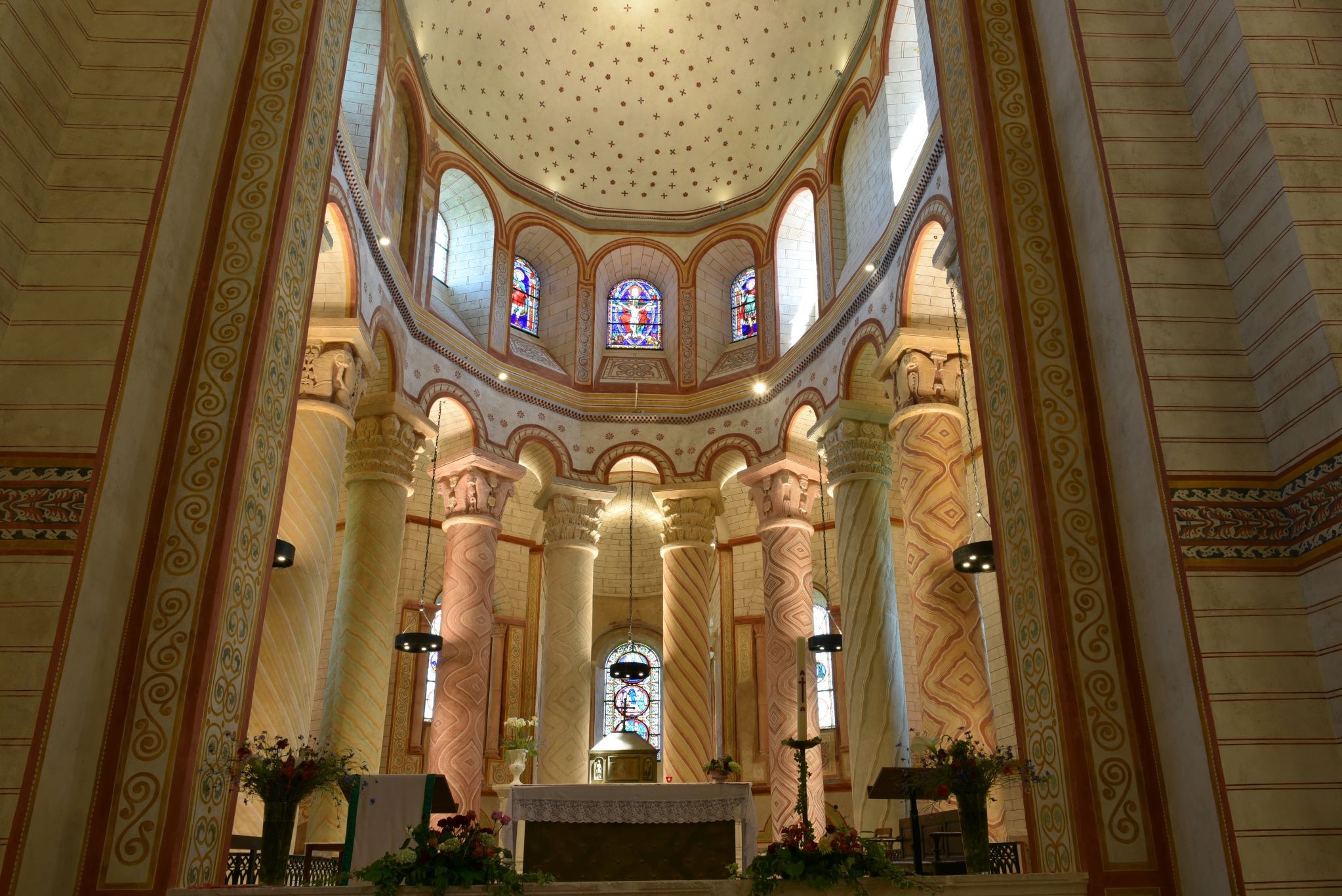 Le coeur de l’abbaye de St Savin, avec son autel au centre avec des bouquets de fleurs et ses pupitres de chaque côté, des lumières suspendus au dessus, et des colonnes peintes qui entourent le coeur éclairée, avec des vitraux au dessus 