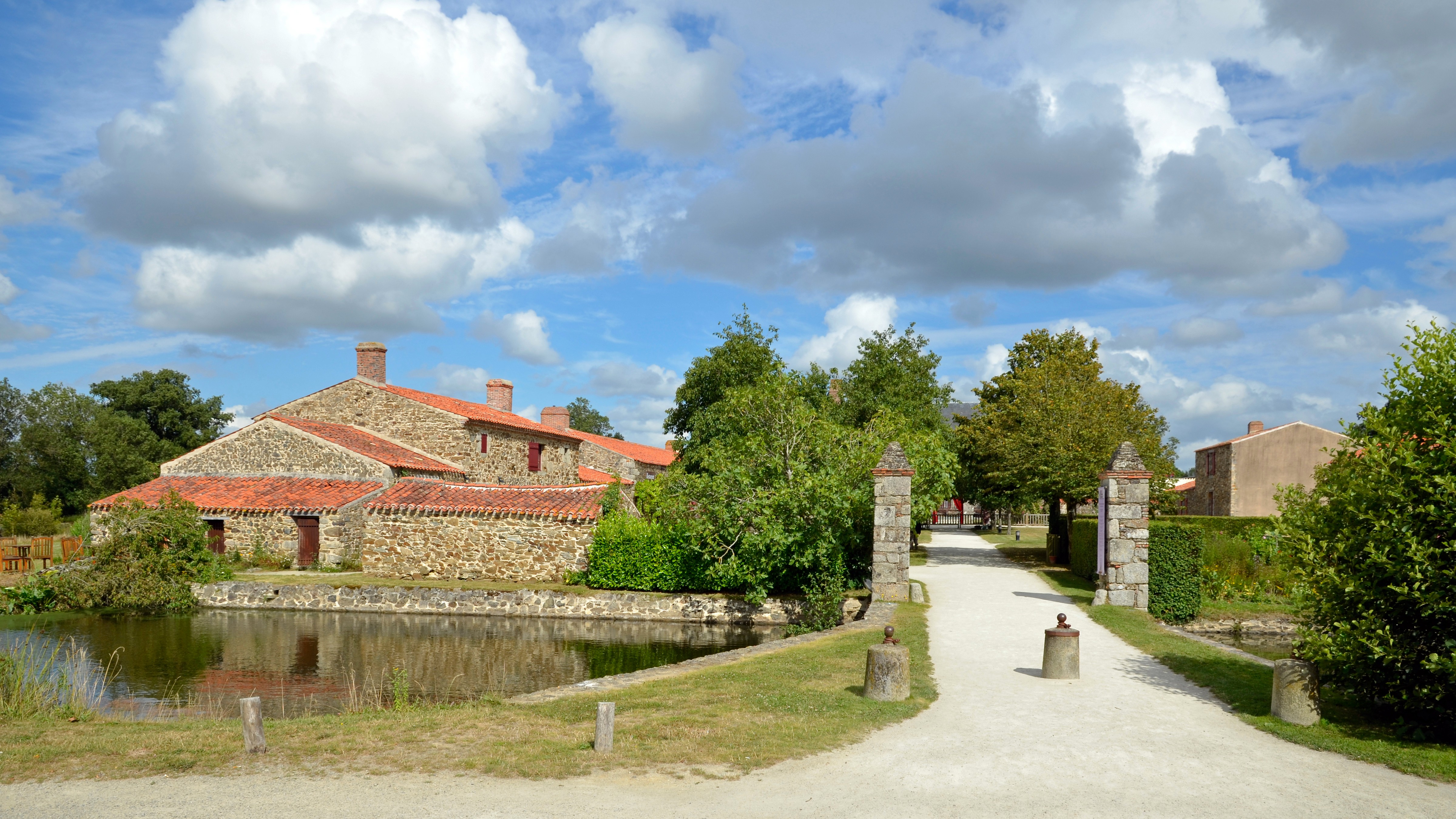 Entrée du domaine avec un accès depuis un pont traversant un cours d’eau et menant à deux piliers à côté du logis en pierre.