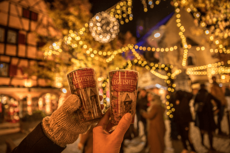 Deux gobelets de vin chaud tenus devant un décor festif à Eguisheim, marché de Noël illuminé.