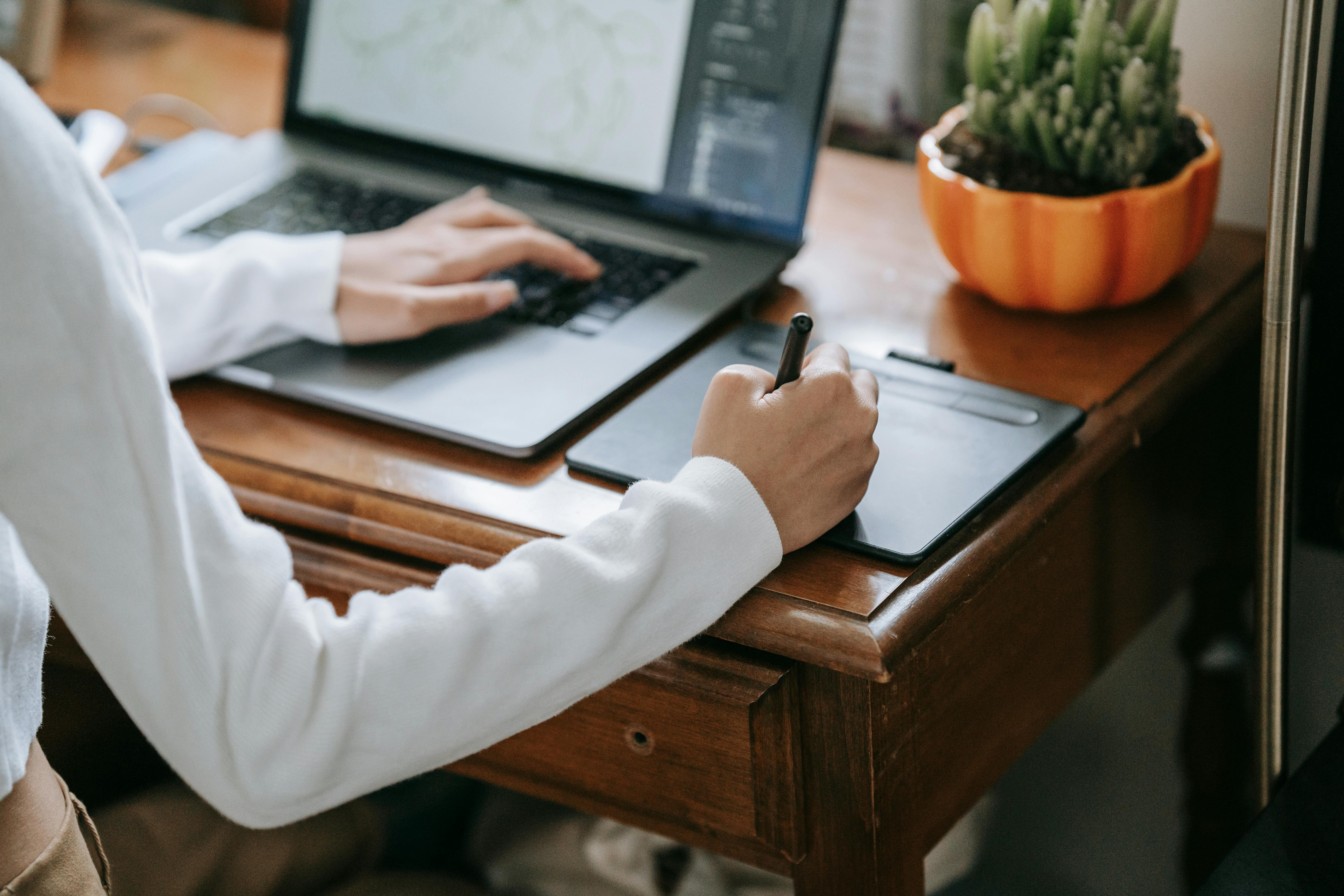 Personne dessinant sur une tablette graphique à côté d'un ordinateur portable, avec une plante en pot en arrière-plan.