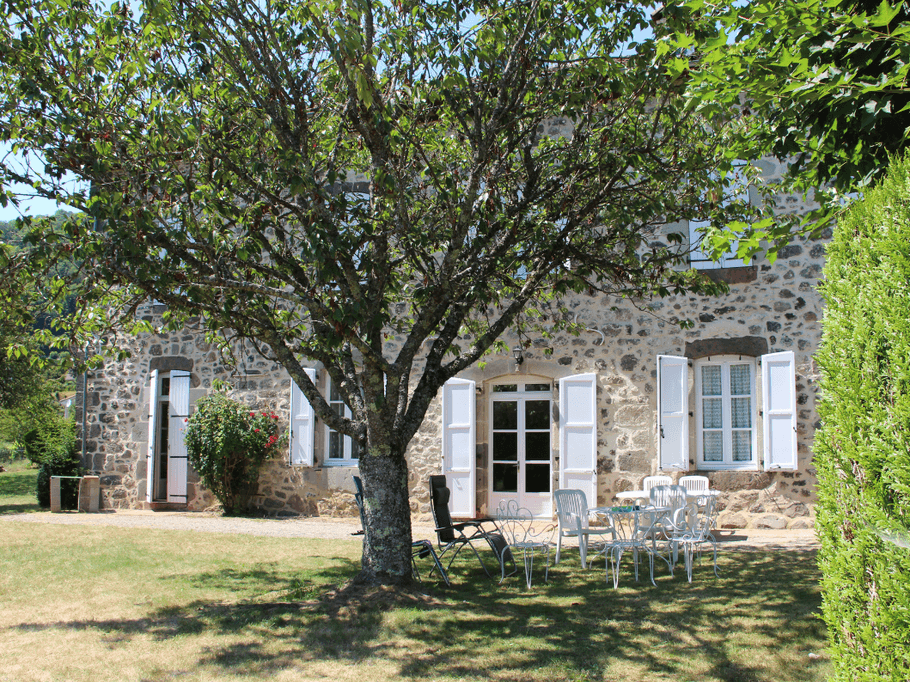 Façade en pierre de la maison Mirabel avec jardin arboré et mobilier d’extérieur, parfaite pour un séjour estival au Cantal