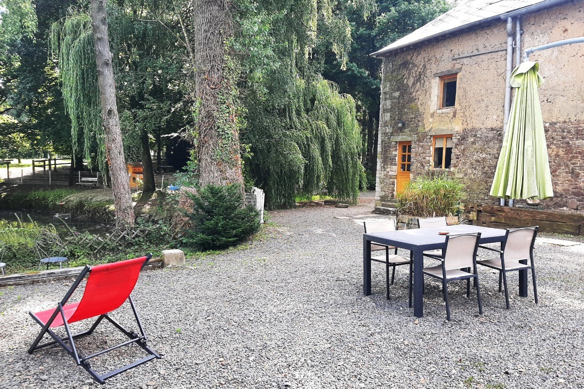 La façade arrière du gîte avec sa cour privative à la location, son salon de jardin, parasol, transats et vue sur l'étang.
