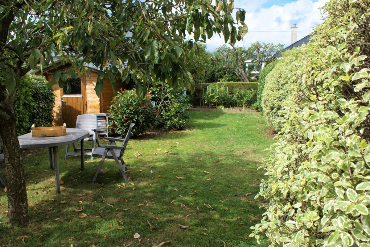 Façade arrière de la maison. Terrasse avec salon de jardin et plancha aux beaux jours. Jardin arboré et stockage vélos.